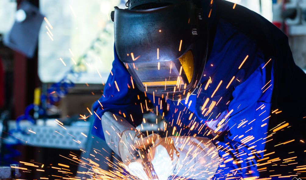 Welder wearing a protective helmet, working with sparks flying from a metalwork welding process in a workshop.